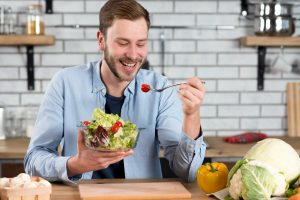 Un homme souriant portant une liquette bleue et un T-Shirt noir, assis dans une cuisine, tenant un grand bol de salade et entrain de regarder sa fourchette avec une tomate.