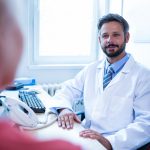 Un médecin portant une blouse blanche assis à son bureau. Devant lui sont posés un téléphone et le clavier d'un ordinateur.